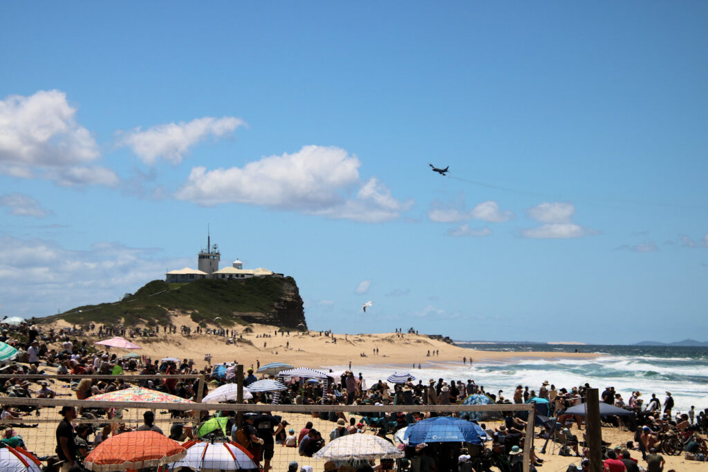 The crowd on the beach while a Lockheed P-3C Orion flies over Nobbys Headland