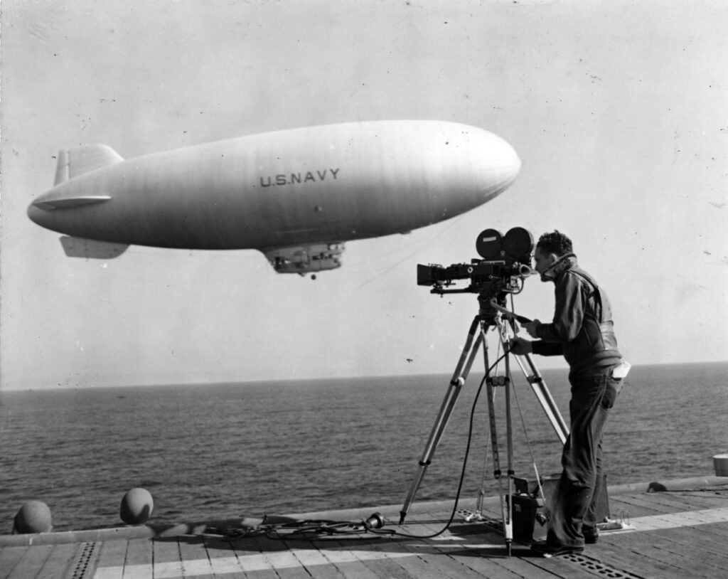 A U.S. Navy blimp looms in the background as a cameraman films from the deck of the carrier USS Matanikau (CVE-101)