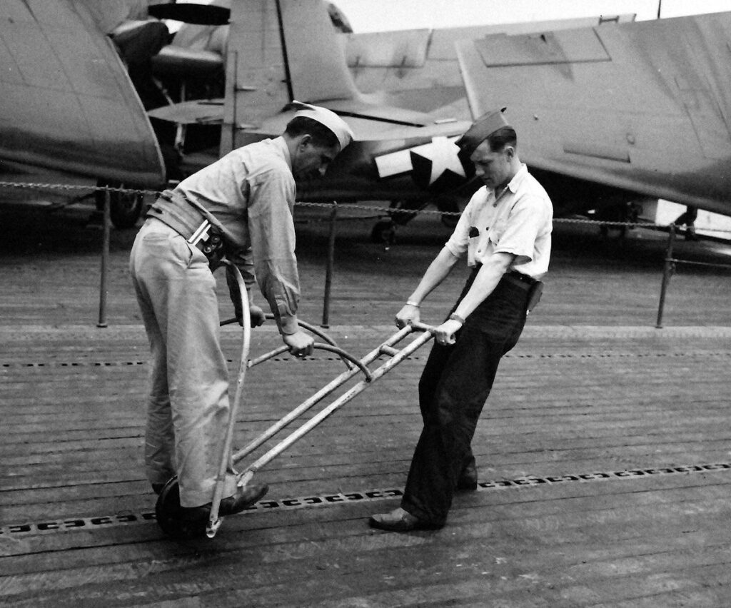 Crewmembers of USS Matanikau (CVE-101) use an implement to sink tar into grooves in the flight deck, 11 August 1944