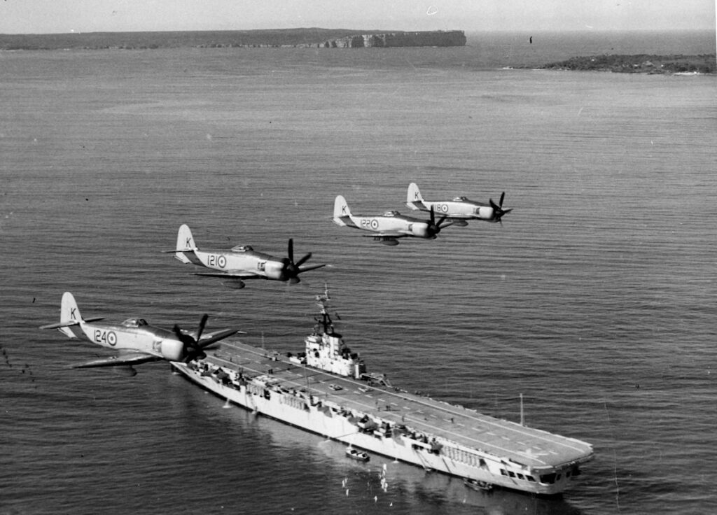 HMAS Sydney in Sydney Harbor. Four Hawker Sea Fury fighter planes fly in formation over the aircraft carrier, circa 1949